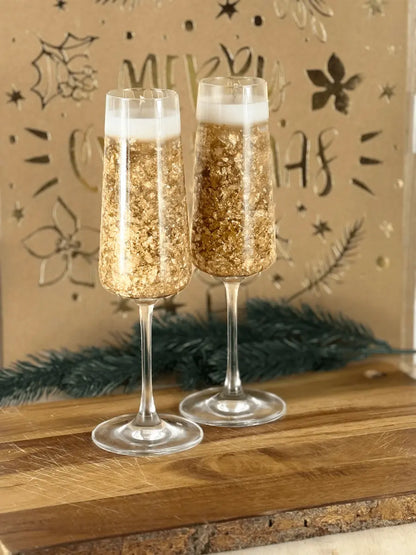 Handgemachte Dessertkerze im Champagnerglas mit goldenen Sprudelelementen auf Holzbrett vor festlicher Deko – kramMITcharme